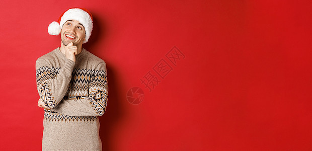 戴着圣诞帽和圣诞毛衣的快乐年轻人的形象 想象着一些东西 想着新年礼物 微笑着 看着左上角 站在红色背景上 快乐的 购物背景图片