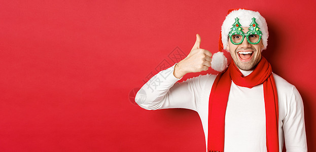 圣诞 冬季假日和庆祝的观念 穿着圣塔帽和派对眼镜的欢快男子 举起拇指并微笑着高兴 站在红色背景之上 毛衣 脸图片