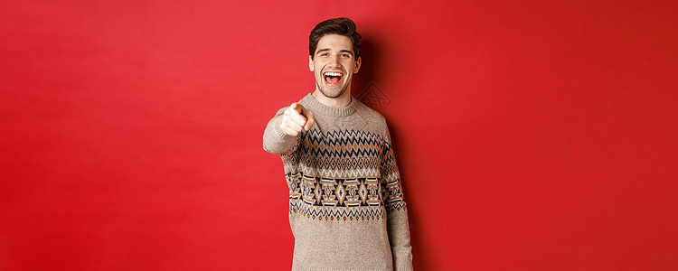 圣诞节庆典 冬季和生活方式的概念 穿着圣诞毛衣的快乐帅哥 祝节日快乐 指着相机 站在红色背景上 微笑 工作室图片