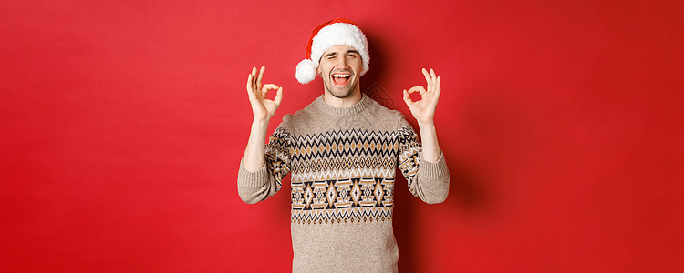 穿着毛衣和圣塔帽的喜悦有魅力的家伙 祝愿圣诞快乐 展示好迹象 对镜头眨眼 庆祝新年 站在红背景上 男人 购物图片