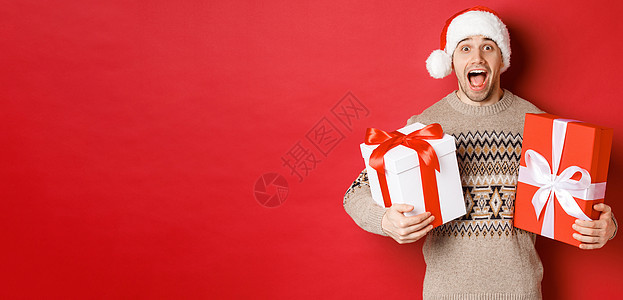 欢快迷人的男人拿着圣诞礼物 戴着圣诞帽和冬日毛衣 微笑着惊讶地站在红色背景上的形象 工作室 成人图片