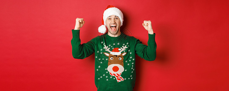 欢快迷人的男人庆祝新年的画像 穿着绿色毛衣和圣诞帽 欢呼雀跃 胜利或胜利 站在红色背景上 美丽的 冬天图片
