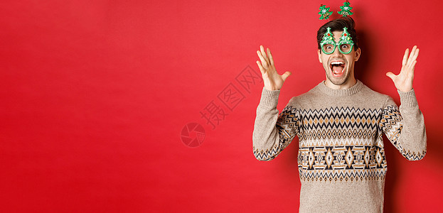 令人兴奋和惊异的男子在派对眼镜 圣诞节毛衣 举起手来大宣布 享受新年庆典 红背景 庆祝 冬天背景图片