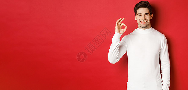 快乐而满意的帅哥的画像 穿着白色毛衣 展示着好的手势 微笑着 赞同一些好东西 推荐商店 站在红色背景上 年轻的 派对图片