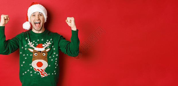 欢快迷人的男人庆祝新年的画像 穿着绿色毛衣和圣诞帽 欢呼雀跃 胜利或胜利 站在红色背景上 男生 成就图片
