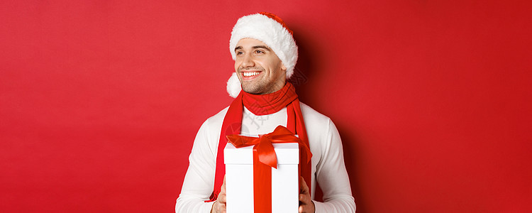 寒假 圣诞节和生活方式的概念 戴着圣诞帽和围巾的帅哥的特写 拿着新年礼物微笑着向左看 站在红色背景上 黑发 男人背景图片