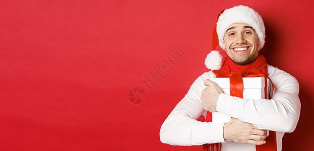 寒假 圣诞节和生活方式的概念 戴着圣诞帽和围巾的可爱男人的形象 拥抱他的新年礼物 微笑着受宠若惊 站在红色背景上 庆祝 圣诞节快图片