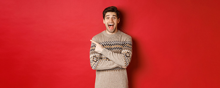 惊艳帅哥的肖像 穿着圣诞毛衣 手指指着左上角的标志 看起来很兴奋 展示了很棒的新年促销优惠 站在红色背景上 大胡子 冬天背景
