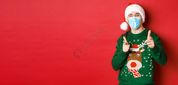 新年 covid-19 和社会疏远的概念 穿着圣诞毛衣 医用面具和圣诞帽的快乐男人 用手指指着镜头 祝节日快乐 站在红色背景上 图片