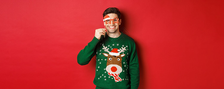 穿着圣诞节毛衣和派对眼镜 庆祝新年 玩乐 站在红色背景下站立的英俊笑脸男子肖像 男人 购物背景图片
