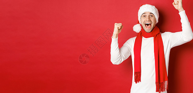 欢乐的男子在圣塔帽和围巾中 呼喊欢乐 举起手 庆祝胜利或胜利 战胜红色背景 冬天图片