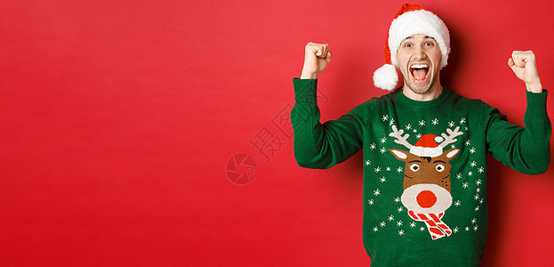 欢快迷人的男人庆祝新年的画像 穿着绿色毛衣和圣诞帽 欢呼雀跃 胜利或胜利 站在红色背景上 快乐图片