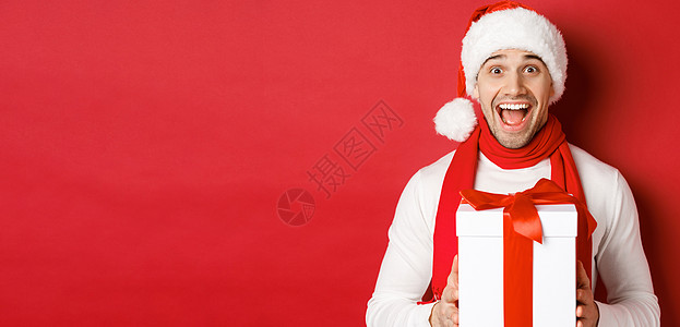 寒假 圣诞节和生活方式的概念 身穿圣诞老人和围巾的帅哥兴奋的特写 看起来很惊讶 收到新年礼物 站在红色背景上 促销 时尚背景图片