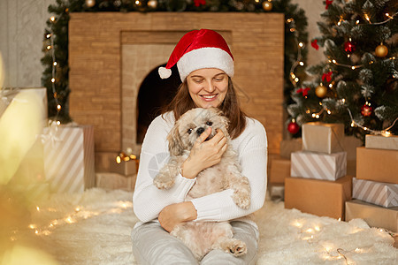 戴着红帽的可爱快乐女孩在圣诞树的背景下与可爱的北京狗拥抱 节日房间里有灯光 壁炉和礼品盒 女性坐在地板上 和小狗一起玩图片