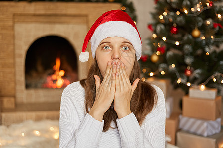 年轻的欧洲妇女佩戴圣塔帽和白色毛衣 在圣诞节装饰院周围在家 用手顶嘴 非常惊讶 背着壁炉和X马树摆姿势图片