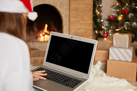照片中迷人的女士坐在地板上 拿着带空白屏幕的笔记本电脑 戴着红色的圣诞老人帽子和白色毛衣 在装饰精美的圣诞客厅里摆姿势 复制空间图片