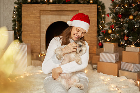 戴着红色帽子和休闲毛衣的女人在圣诞节背景下抱着滑稽可爱的小狗 带着露齿微笑的女士坐在柔软的地毯上 开心地笑着图片