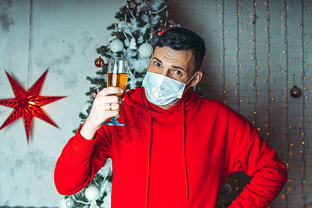 在圣诞树的背景下 戴着医用面具的年轻人拿着一杯香槟 在冠状病毒大流行期间庆祝安全圣诞节的成年男性 酒精 暴发图片