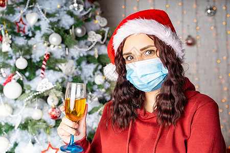 圣诞树背景下 戴着医用面具 头戴圣诞帽 端着香槟的年轻女子 戴着防护面具的迷人黑发女郎在冠状病毒大流行期间庆祝安全的圣诞节 享受图片