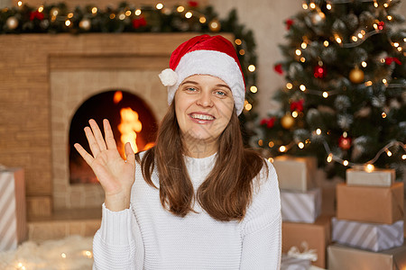 年轻女子快乐而愉快地微笑着 挥手欢迎和问候某人或说再见 戴着圣诞老人的帽子和毛衣 在圣诞树 壁炉和装有礼物的盒子附近摆姿势图片