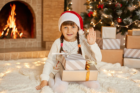 快乐的小女孩在平安夜坐在地板上的圣诞树附近 对着相机挥手 摆着一堆礼品盒 孩子穿着休闲服和红色圣诞帽图片