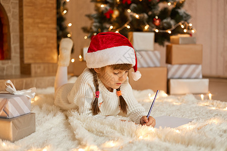 可爱的小女孩在家里写信给圣诞老人 靠近礼品盒 躺在柔软的地毯上 扎着辫子的孩子在圣诞树旁摆姿势图片