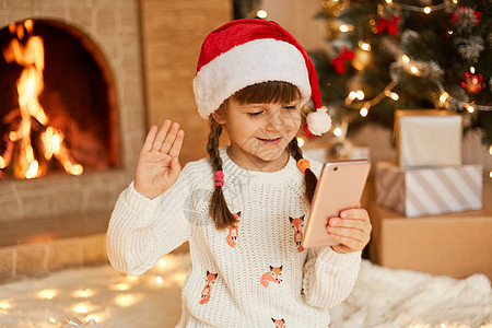 快乐的孩子看着智能手机屏幕 挥舞着手 给朋友或亲戚打电话 在家里过圣诞节 穿着白色毛衣 戴着喜庆的圣诞老人帽 靠近壁炉图片