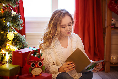 坐在圣诞树旁边的地板上 并阅读圣诞节明信片的年轻美少女背景图片