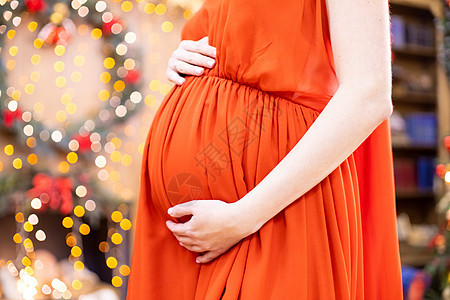 最好的圣诞礼物 身穿红色连衣裙的孕妇在模糊的圣诞客厅背景下抱住她的肚子 特写没有脸 怀孕 健康图片