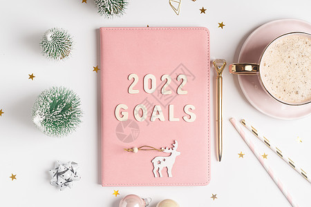 顶部视图桌面圣诞粉色记事本 带有 2021goals 字母文本 平铺的白桌背景 配有规划师 咖啡 圣诞装饰 笔记本和文具 金的 图片