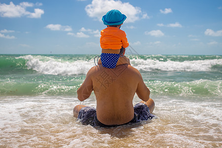 家庭价值观概念 爸爸后卫 宝宝坐在爸爸的肩膀上 爸爸坐在沙滩上 海浪来了图片