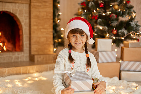 家里地板上戴着红色帽子的可爱女孩 用礼物庆祝圣诞 小女孩拿着礼盒摆姿势 对着镜头微笑 穿着白色套头衫 坐在壁炉和圣诞树旁图片