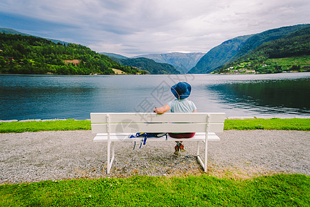 坐在长椅上看着挪威乌尔维克峡湾的女人 挪威霍达兰县乌尔维克的峡湾海滨长廊 戴帽子的孤独游客坐在长凳上欣赏斯堪的纳维亚的风景 夏天图片