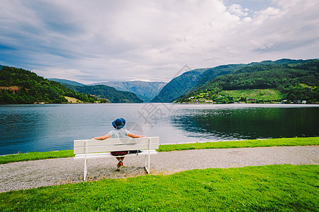 坐在长椅上看着挪威乌尔维克峡湾的女人 挪威霍达兰县乌尔维克的峡湾海滨长廊 戴帽子的孤独游客坐在长凳上欣赏斯堪的纳维亚的风景 旅游图片