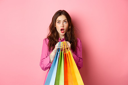 女人被打折扣震惊 用惊异的脸仰望起来 拿着购物袋 站在粉红背景上站立 女士 快乐的图片