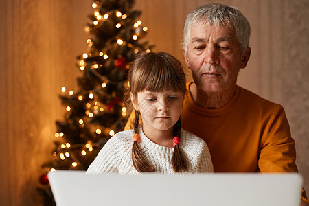 身穿橙色毛衣的成年男子和坐在笔记本电脑前 背着圣诞树的小女孩在门内拍摄 他们以高度集中的表情观看监视器图片
