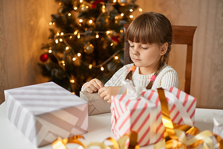 小女孩扎着两条辫子坐在桌旁 打包圣诞盒 背景是圣诞树 孩子神情专注 给家人惊喜图片