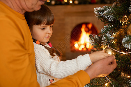 有女儿的不明男子站在Xmas树旁 用Xmas球装饰它 看着Garland的灯光 在背景上布置着模糊的壁炉图片