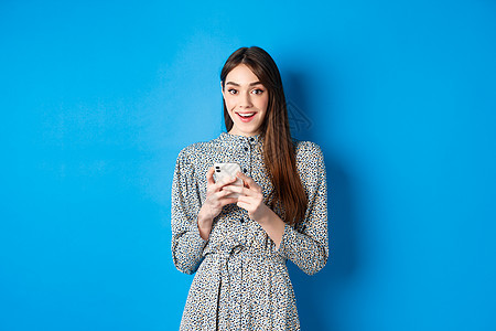 长长的自然头发 穿着衣服 使用智能手机和看起来快乐 站在蓝背景下站立的可爱笑脸女孩 女士图片