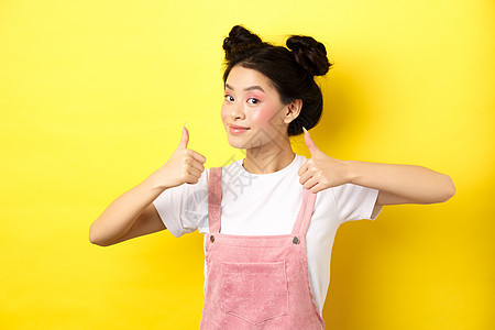 美丽的亚洲女孩 有明亮的粉红色化妆品 露出拇指在认可和笑笑中满足 推荐promo 黄色背景 可爱的图片