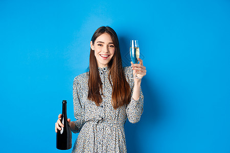 欢庆 快乐的年轻女子穿着洋装 举香槟杯 拿着酒瓶和微笑 在派对上玩乐 站在蓝背景 美丽 投标图片