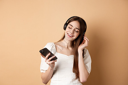 快乐女孩在耳机和微笑中聆听音乐 手握智能手机 蜜蜂背景 浅褐色的 电话图片