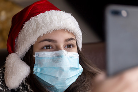 一位身戴圣达克萨斯帽子的年轻女孩 并使用智能手机进行医疗面具谈话 用视频电话给朋友和家长 Corona病毒期间的圣诞节 女性 庆背景图片