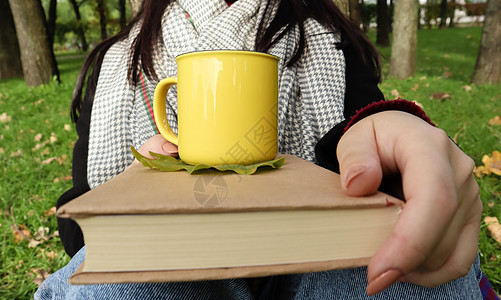 一个穿着外套和围巾的女孩在秋天的森林里拿着一本书和一个杯子 手里拿着一杯热饮 在温暖的一天在城市公园里特写 阅读 放松和舒适的概图片