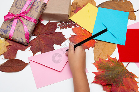 孩子们的手把卡片放在信封里 鲜艳的颜色 带漂亮丝带的礼物 节日问候 感恩节 生日 白桌上的秋叶 复制空间顶视图 粉色 黄色 蓝色图片