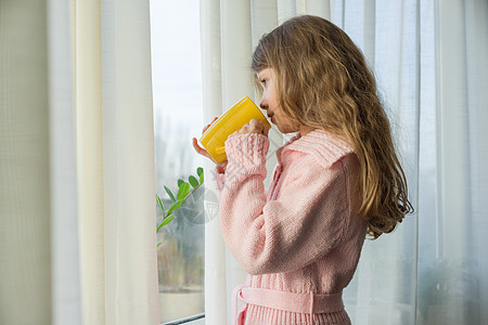 7岁的金发女孩 长长的卷毛 穿着温暖的毛衣 拿着茶杯和窗外看图片