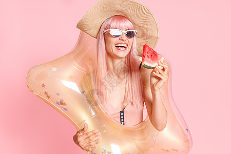 穿着泳衣粉色头发和太阳镜 西瓜切片和漂浮在粉红色背景上游泳的充气星的热辣妇女图片