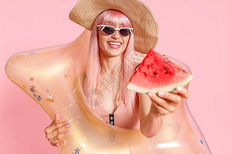 穿着泳衣和太阳镜的粉色头发 带有西瓜片和在粉红色背景上孤立游泳的可充气恒星的兴奋女性图片