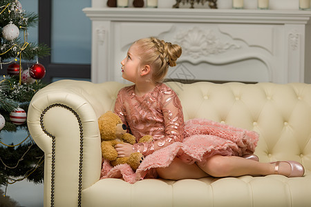 一个小姑娘带着泰迪熊坐在 圣诞树附近的沙发上 玩具 快乐的图片