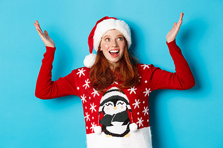 节日快乐和圣诞节的概念 兴奋的红头发女孩为落在她身上的东西而高兴 举起双手高兴地微笑着 戴着带圣诞毛衣的圣诞帽 蓝色背景 冬天 图片